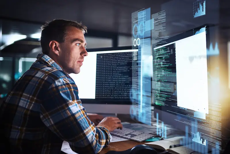 Développeur analysant du code et des données sur plusieurs écrans en environnement professionnel de nuit.