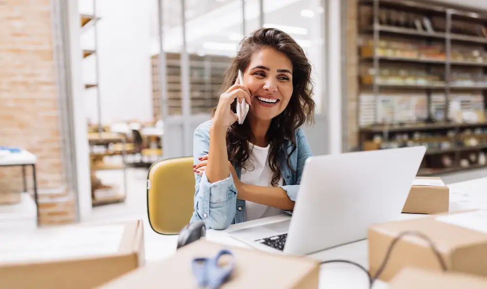 Femme souriante au téléphone travaillant sur son ordinateur portable dans un entrepôt de stockage avec des cartons et des étagères en arrière-plan.