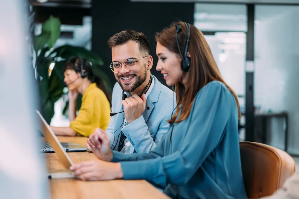 Équipe de support client souriante portant des casques, travaillant ensemble à un bureau dans un centre d'appels moderne.