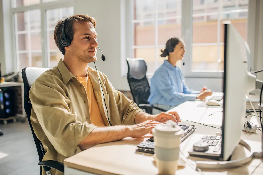 Agent du service client avec casque, assis à son bureau dans un centre d'appels moderne, assistant un client en ligne.